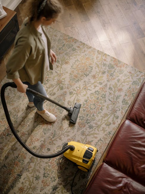 A woman with brown hair tied back, dressed in a light blue, vertically striped shirt and jeans, is kneeling on a patterned rug in a living room, cleaning the dark wooden floor with a blue cloth. The r