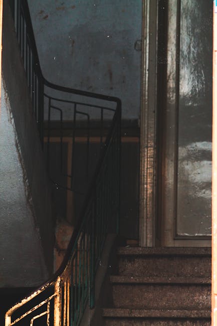 A narrow stairwell with dark, worn concrete steps leading down to a lower floor, enclosed by a high, textured cement wall painted in a muted grey tone. The staircase features a black metal railing wit