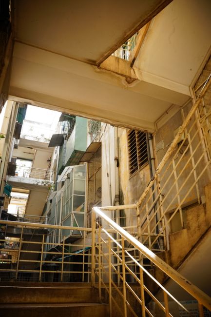 A narrow stairwell with dark, worn concrete steps leading down to a lower floor, enclosed by a high, textured cement wall painted in a muted grey tone. The staircase features a black metal railing wit