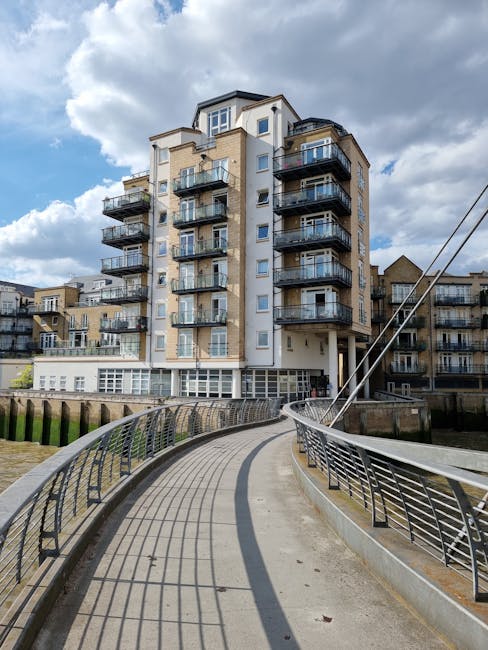 A view of Hammersmith Bridge spanning the River Thames under a cloudy sky, with modern residential buildings along the riverside. The bridge features white-painted suspension cables and towers, partia